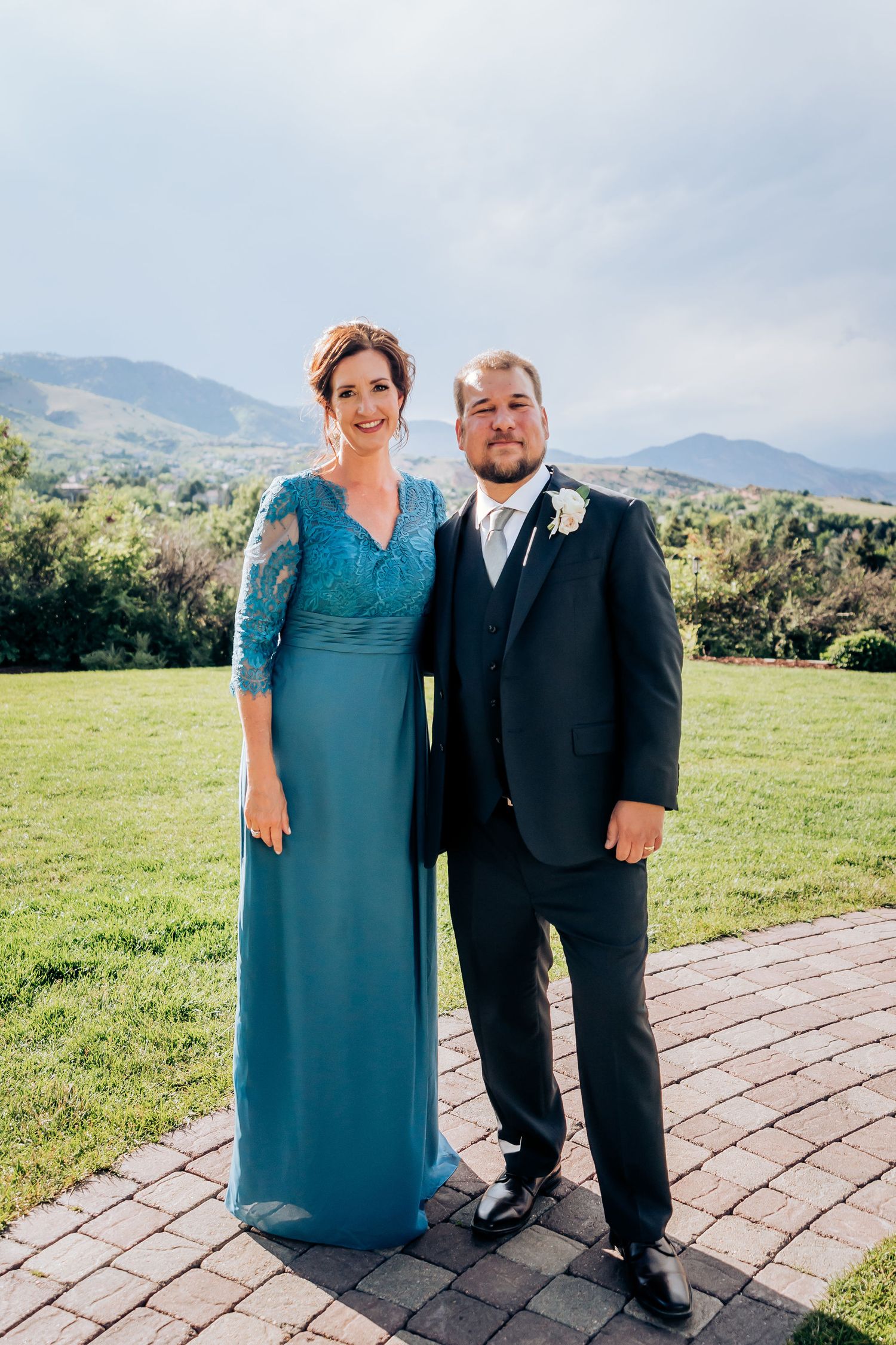 Formal wedding attire portrait featuring teal dress and black suit against mountain landscape.