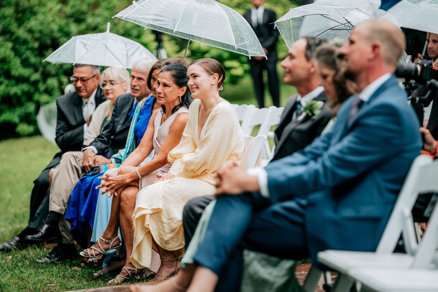 Wedding guests with umbrellas seated in white chairs during outdoor ceremony.