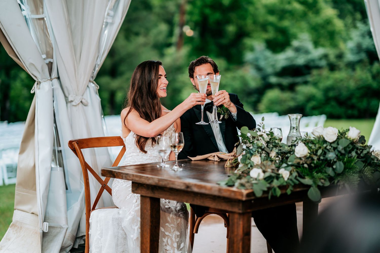 Couple toasts at sweetheart table decorated with greenery under white tent.