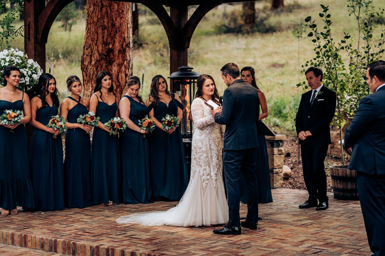 Wedding ceremony taking place on brick platform under wooden pergola surrounded by tall trees.
