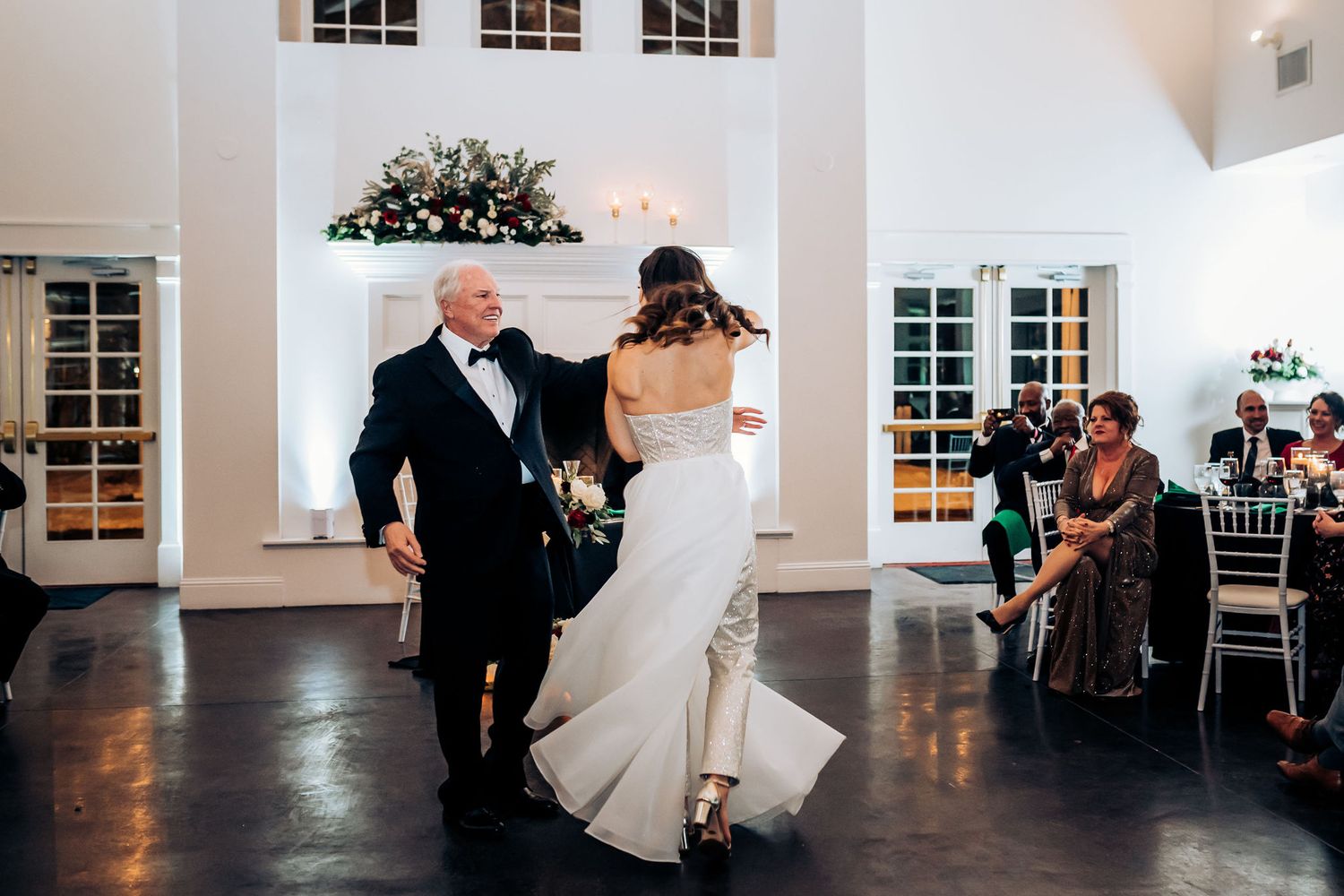 A couple dances together in an elegant white venue with high windows and decorative lighting.