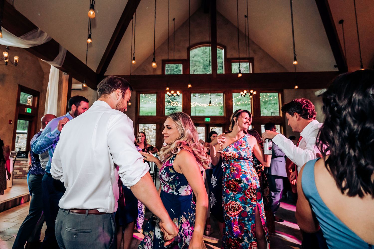 Wedding guests dance and celebrate in a rustic venue with vaulted ceilings and large windows.