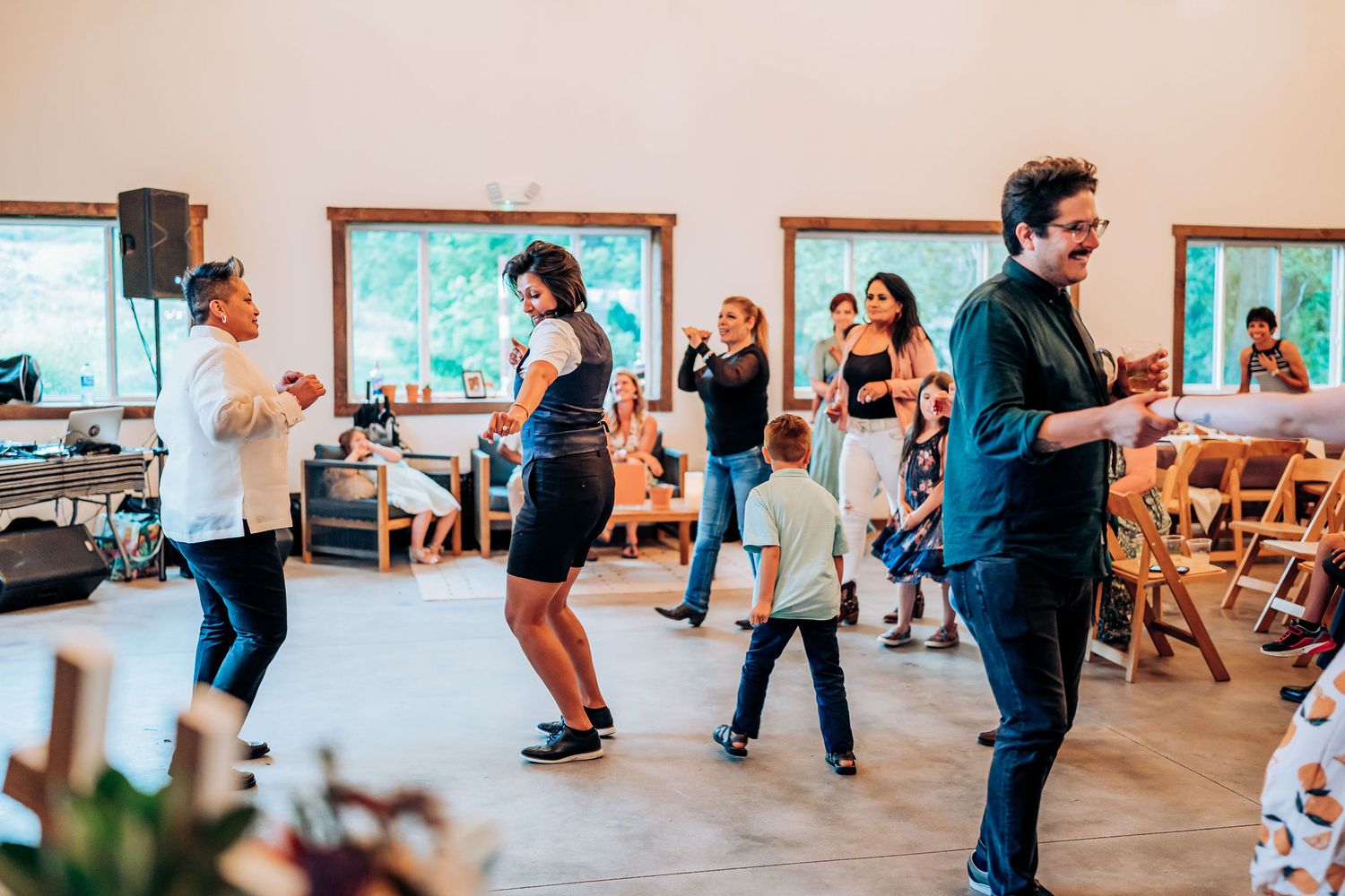 Guests dancing and socializing in a rustic venue with wooden chairs and large windows.