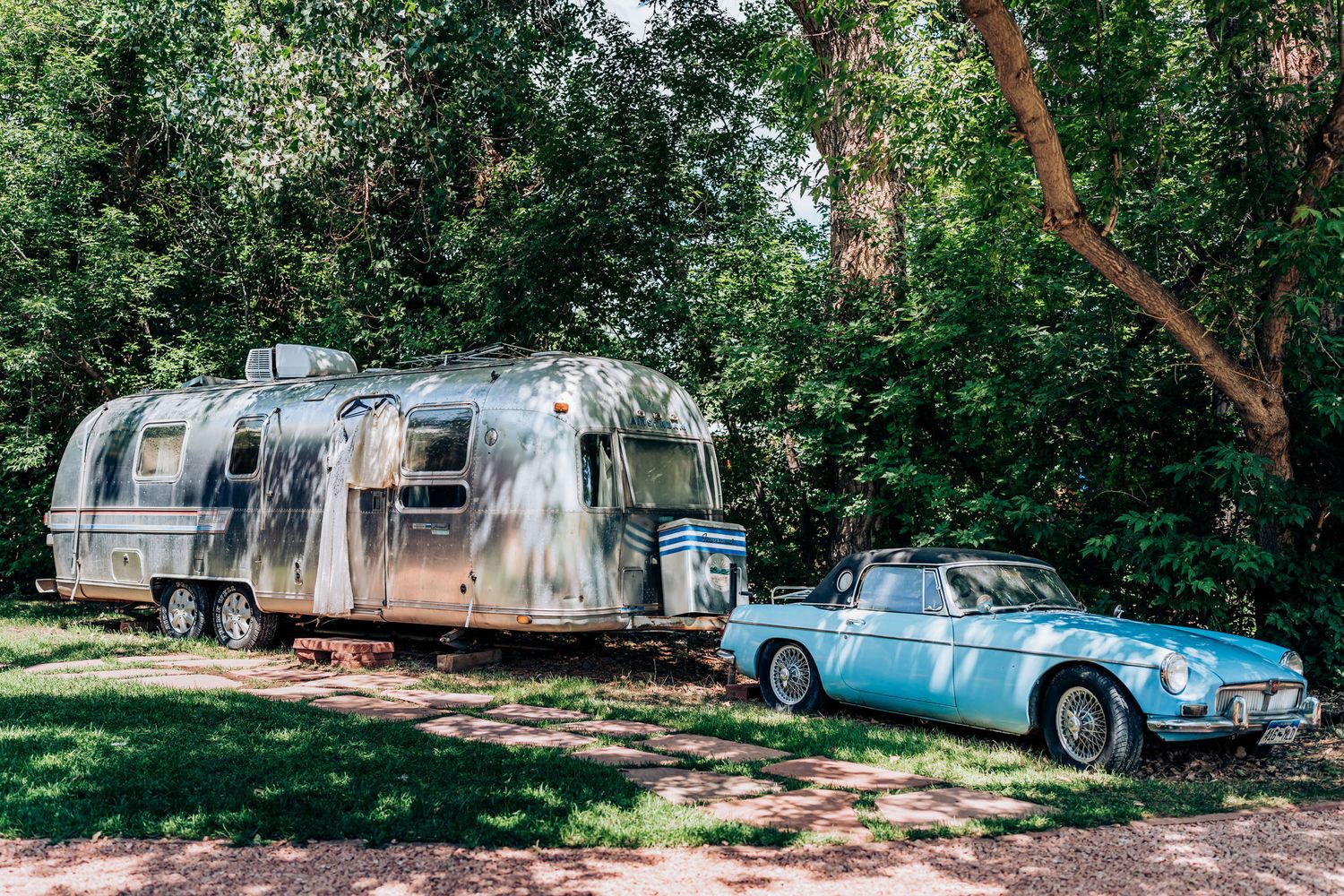 A vintage Airstream trailer parked next to a classic blue car in a wooded setting.