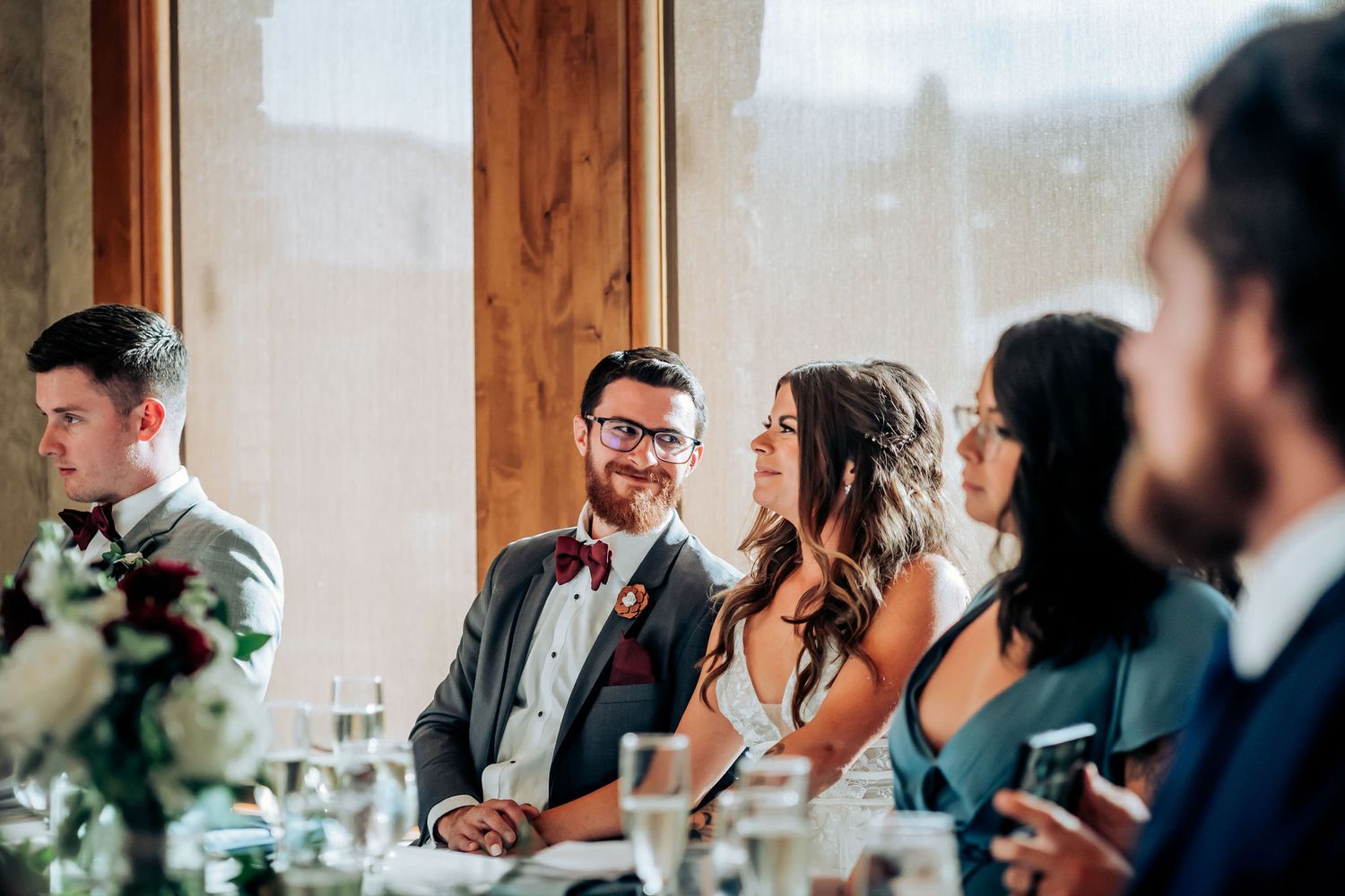 Wedding reception guests smile and laugh together at an elegantly decorated head table with greenery.