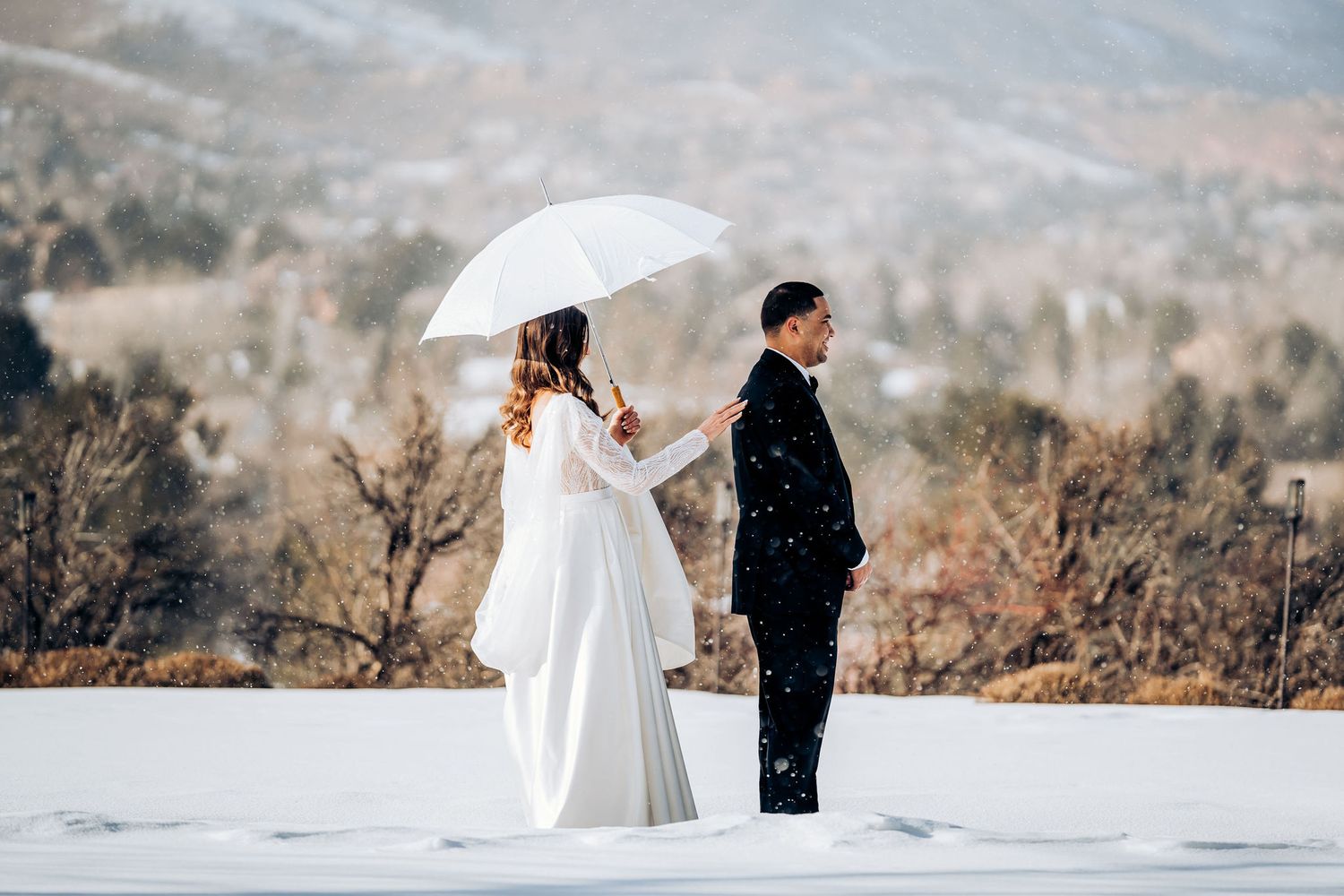 A couple in wedding attire holds a white umbrella while standing in deep snow with mountains behind them.