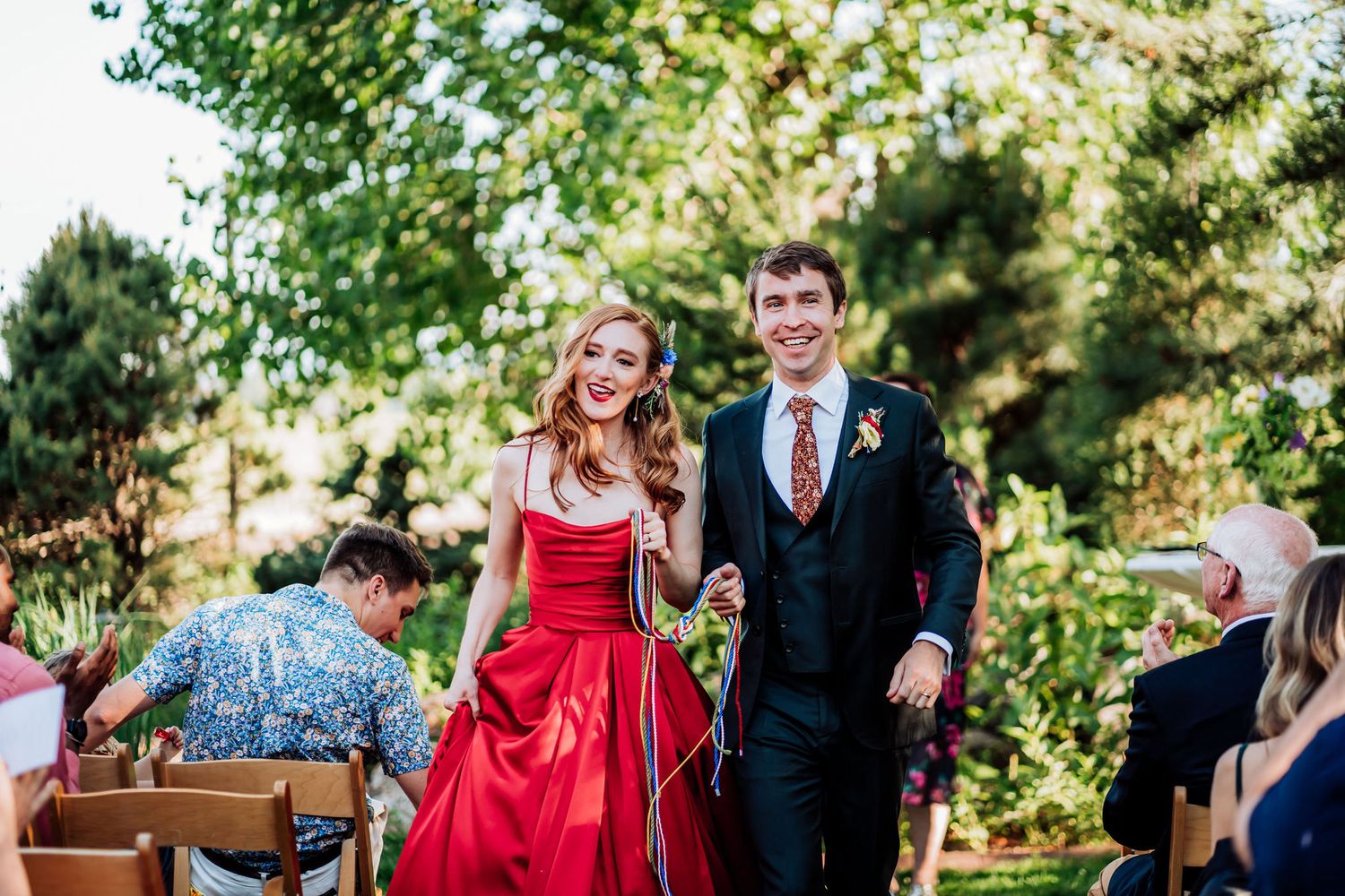 A couple walks down an outdoor aisle together, with the woman in a striking red formal gown.