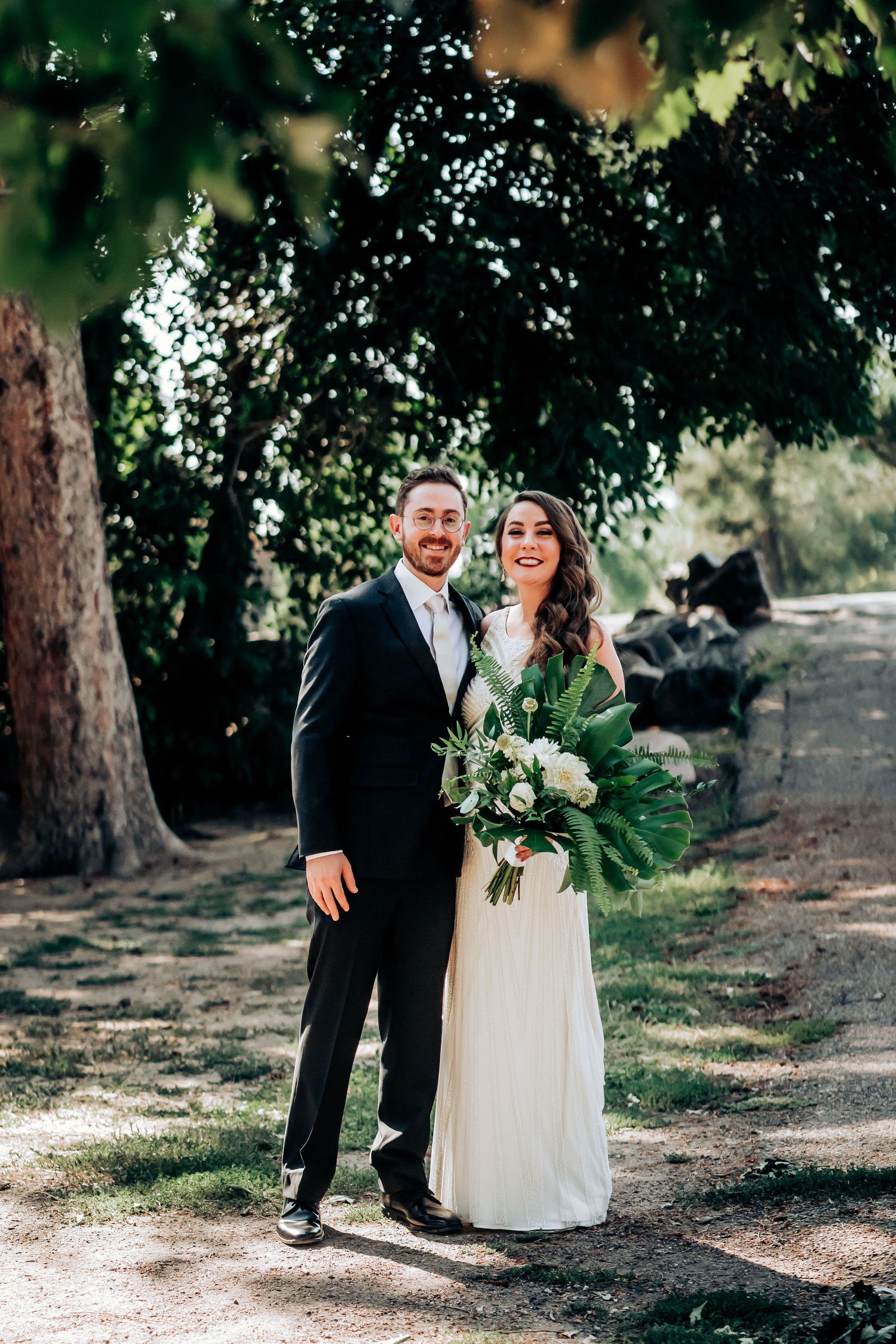A newlywed couple poses with a lush green bouquet in an outdoor garden setting.