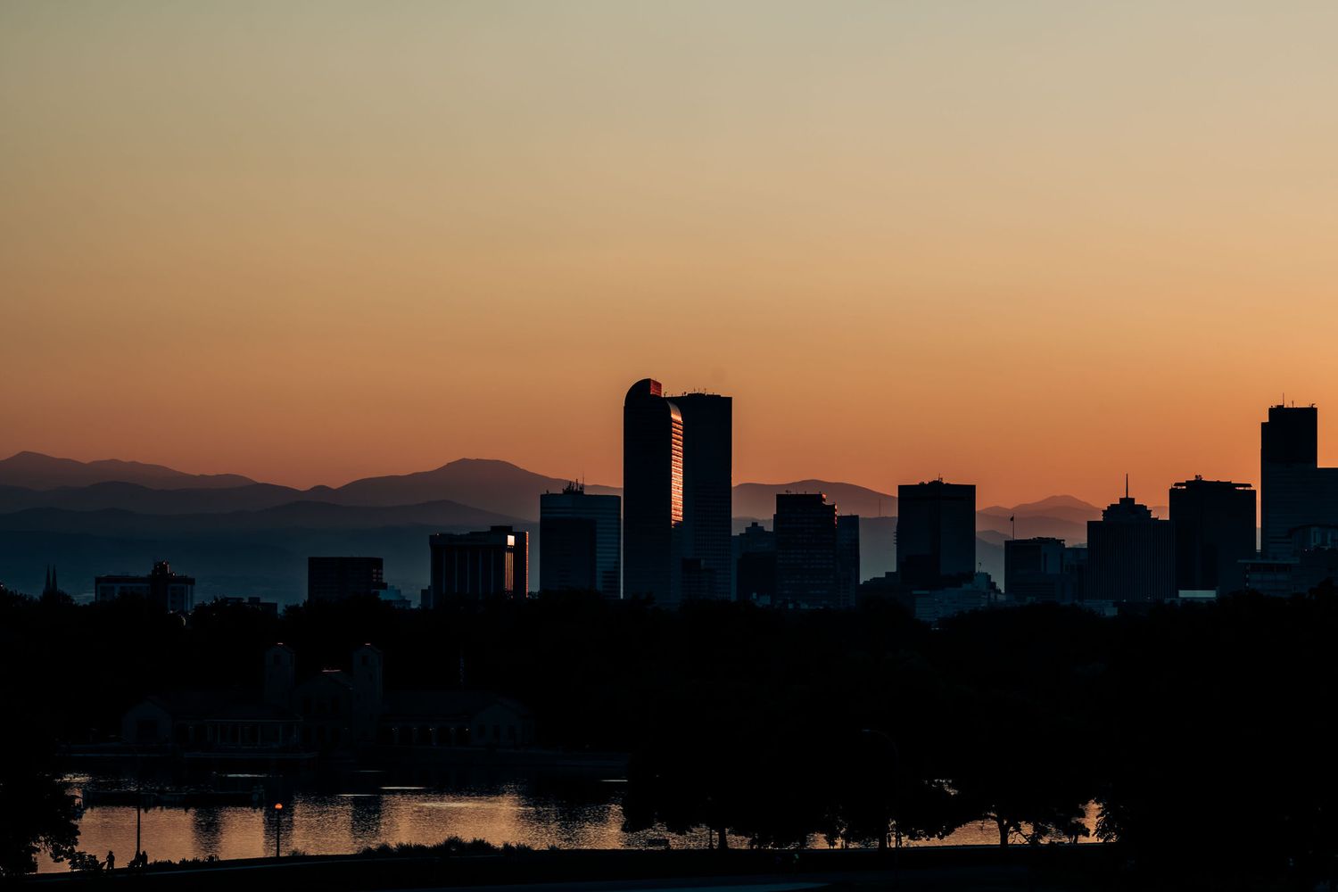 City skyline silhouetted against orange sunset sky with tall buildings and mountains.