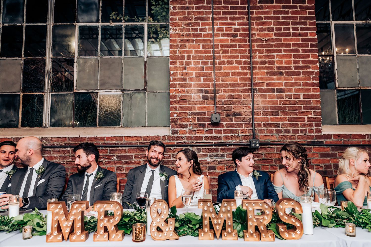 Wedding party seated at head table with 'MR & MRS' letters and greenery decorations against brick backdrop.
