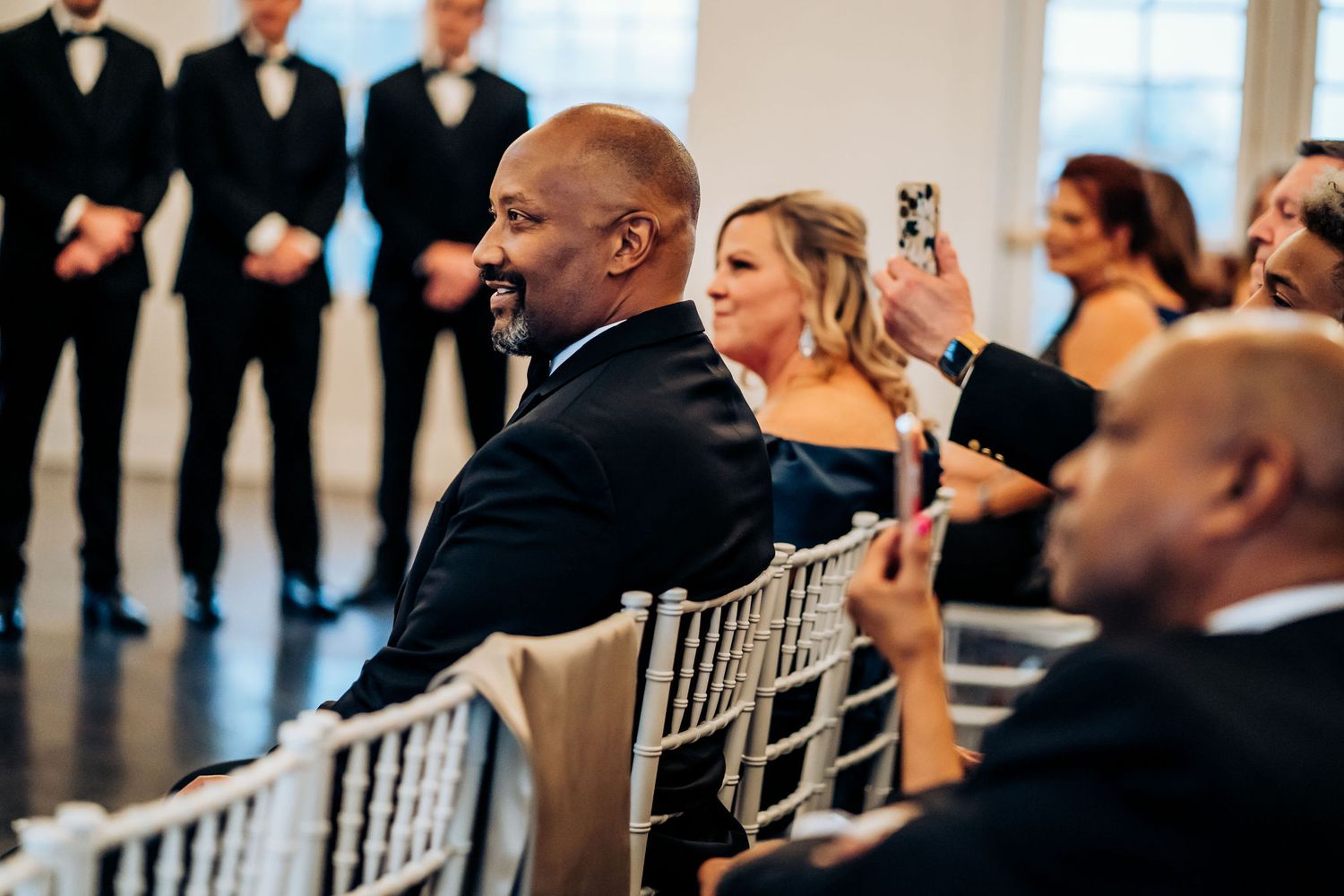 Wedding guests and attendees seated in elegant white chairs smile and take photos during the ceremony.