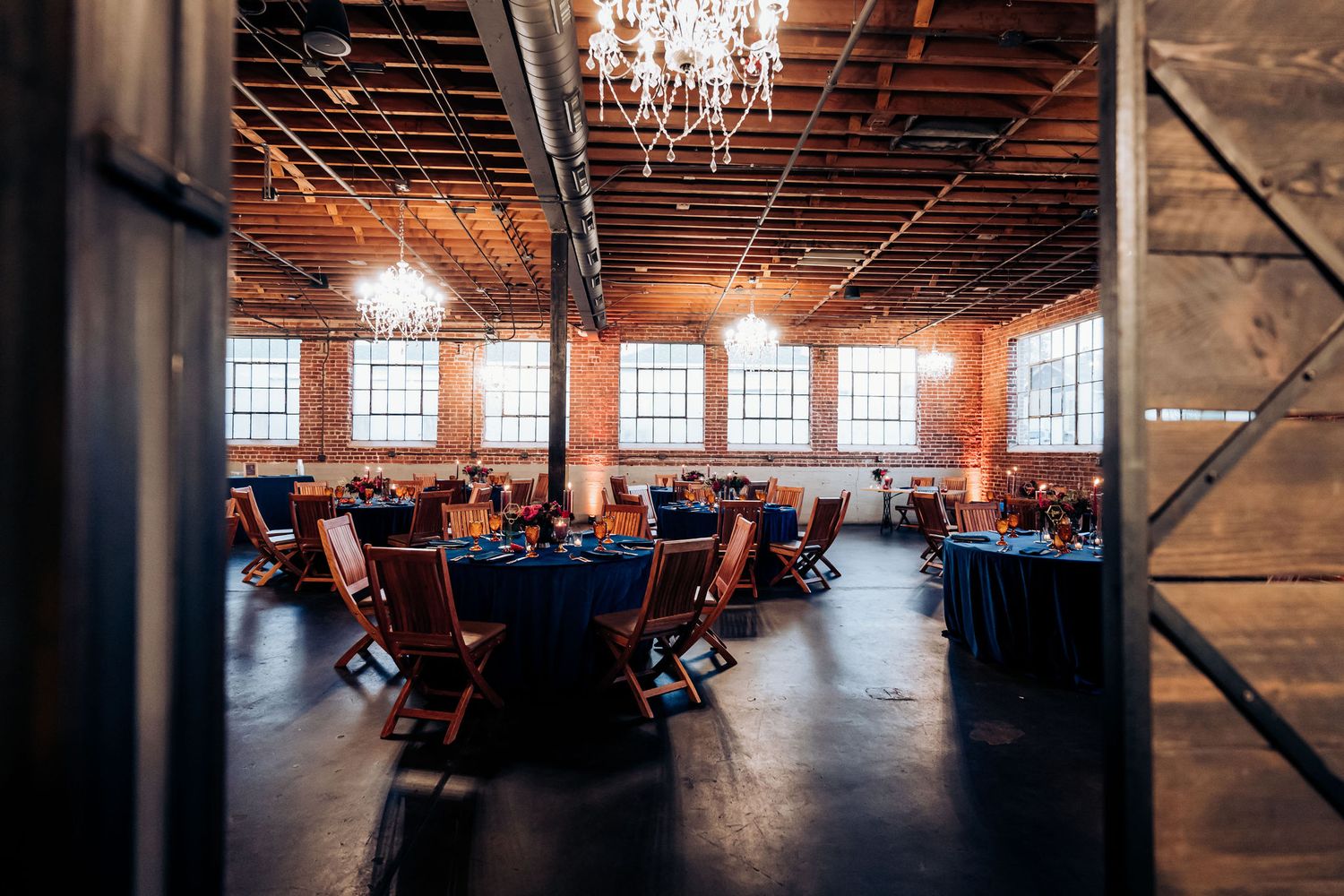 Industrial wedding venue with exposed brick, crystal chandeliers, and navy blue round tables.