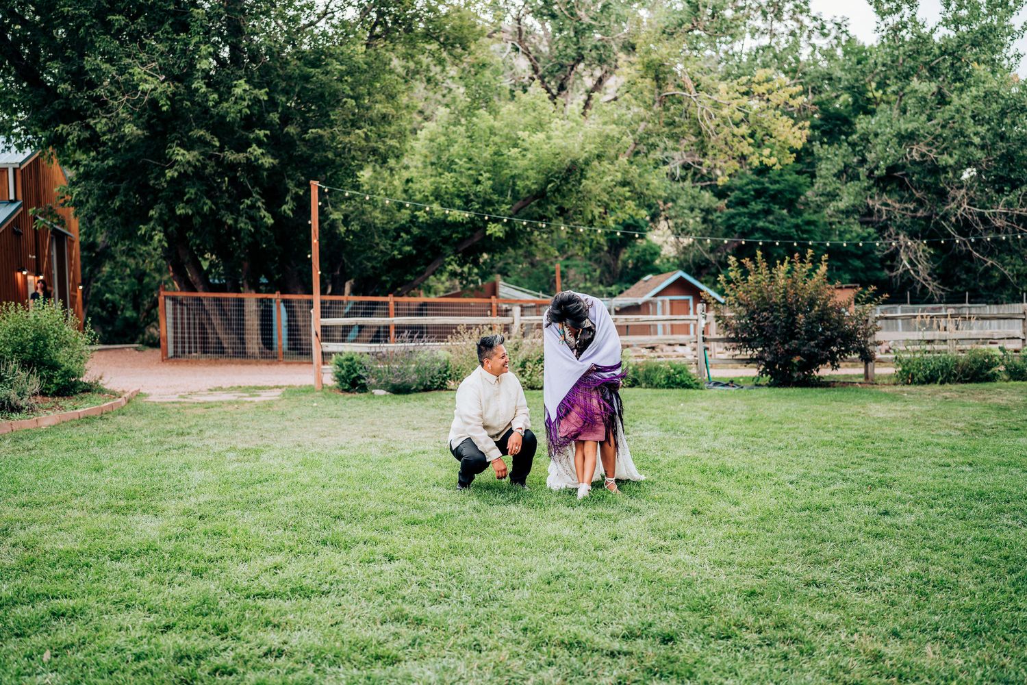 Two figures interact on a grassy lawn with wooden fencing and garden structures in the background.