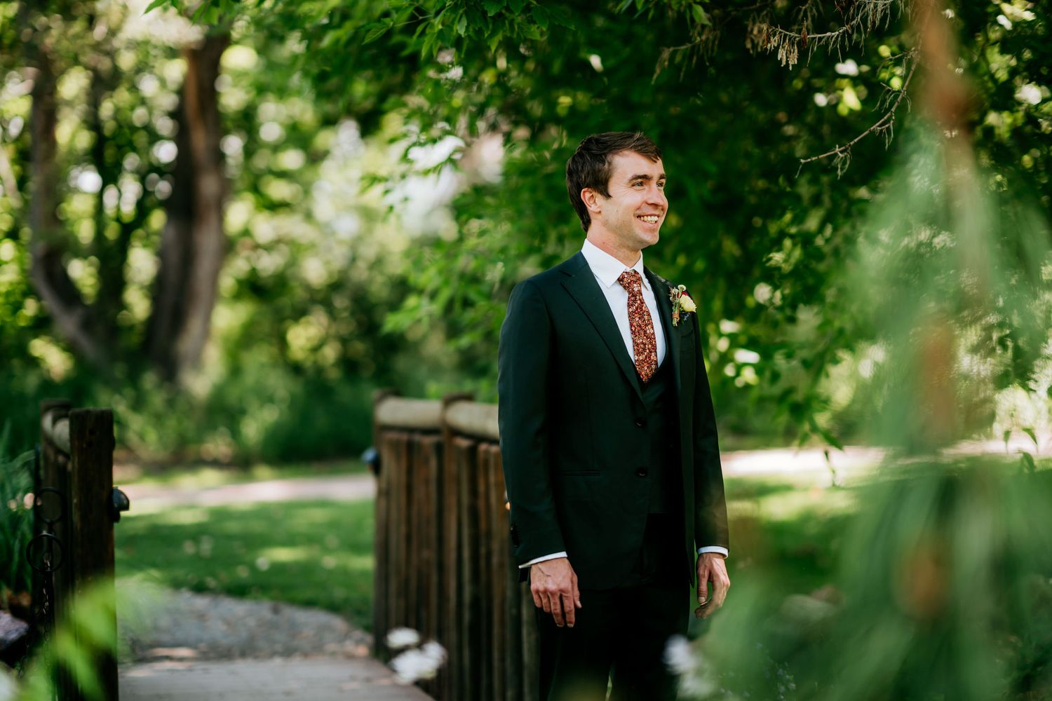 A person in a dark suit stands on a wooden bridge surrounded by lush green foliage.