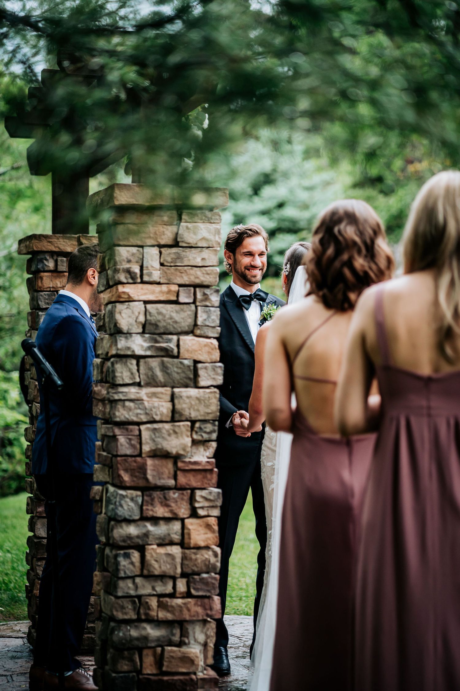 Outdoor wedding ceremony with wedding party members and stone pillars framing the scene.