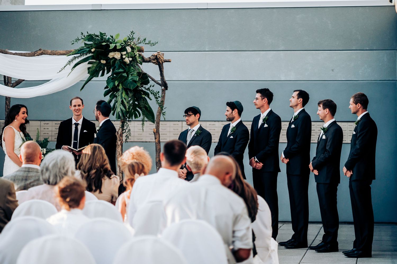 Wedding ceremony featuring groomsmen lined up in black suits against modern backdrop.