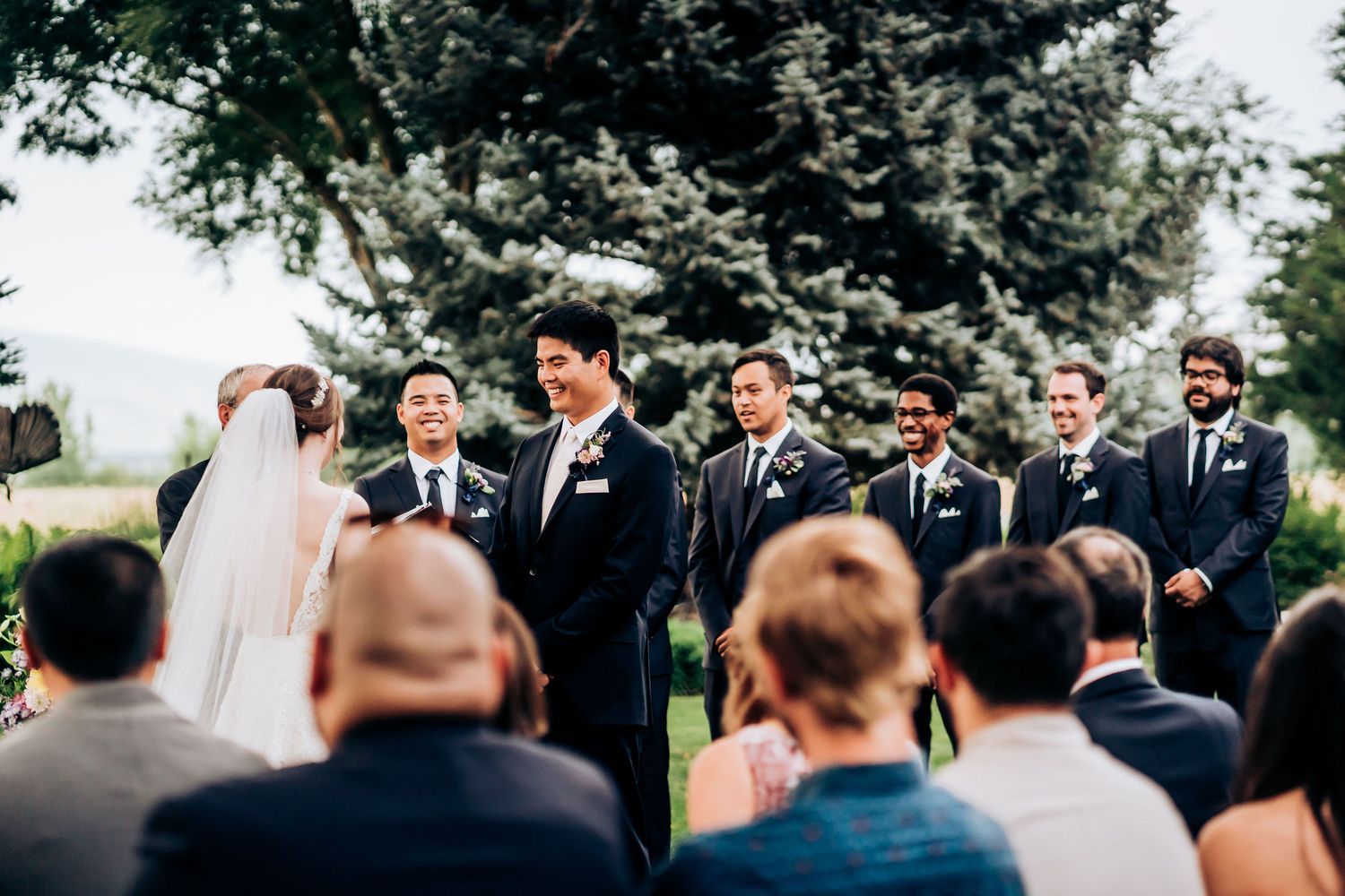Wedding ceremony under towering evergreen trees with guests seated on white chairs.