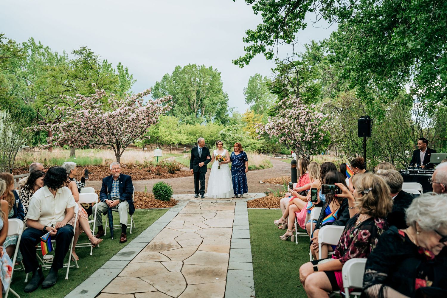 Outdoor garden wedding ceremony with guests seated on white chairs under cherry blossoms.