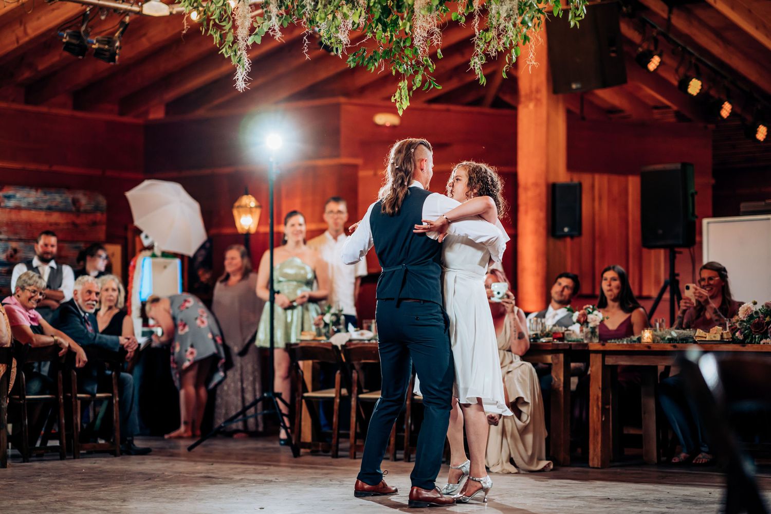 Couple dances in spotlight at reception while guests watch from tables in background.