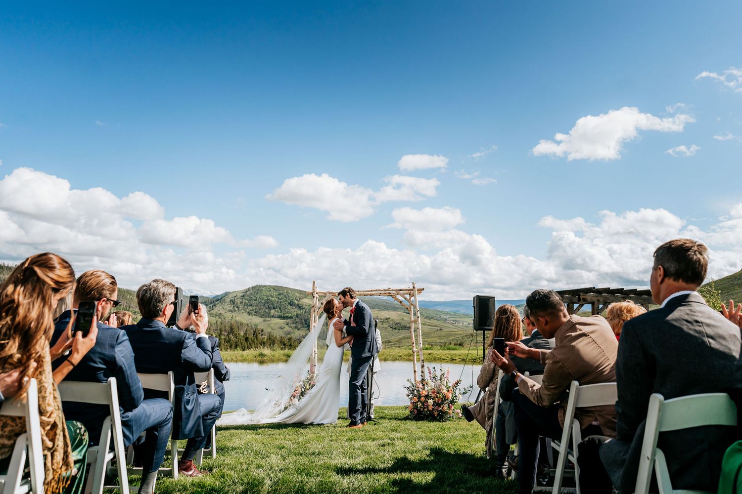 Wedding guests seated on white chairs watching ceremony with mountain landscape in background.