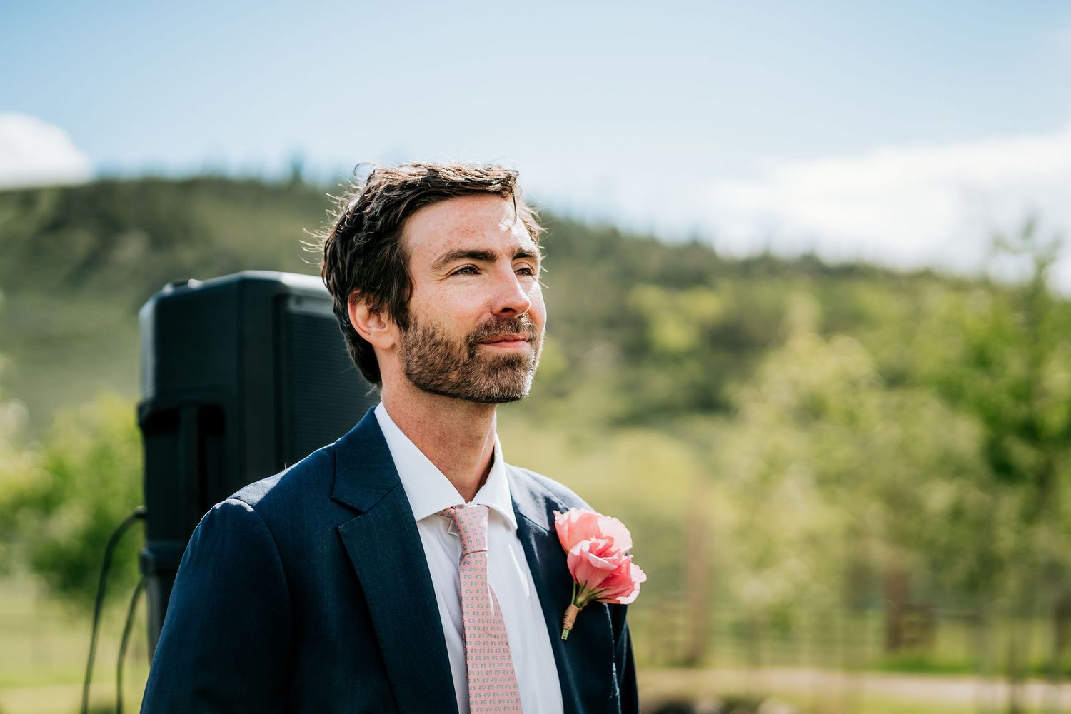 Groom in navy suit with pink boutonniere stands outdoors against mountain backdrop.
