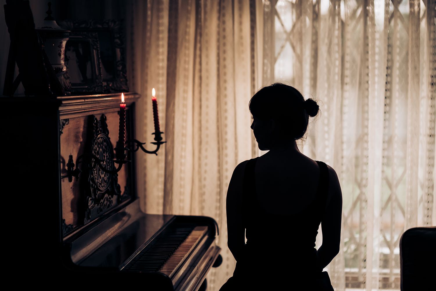 Silhouette near a piano and candlestick against curtained windows.