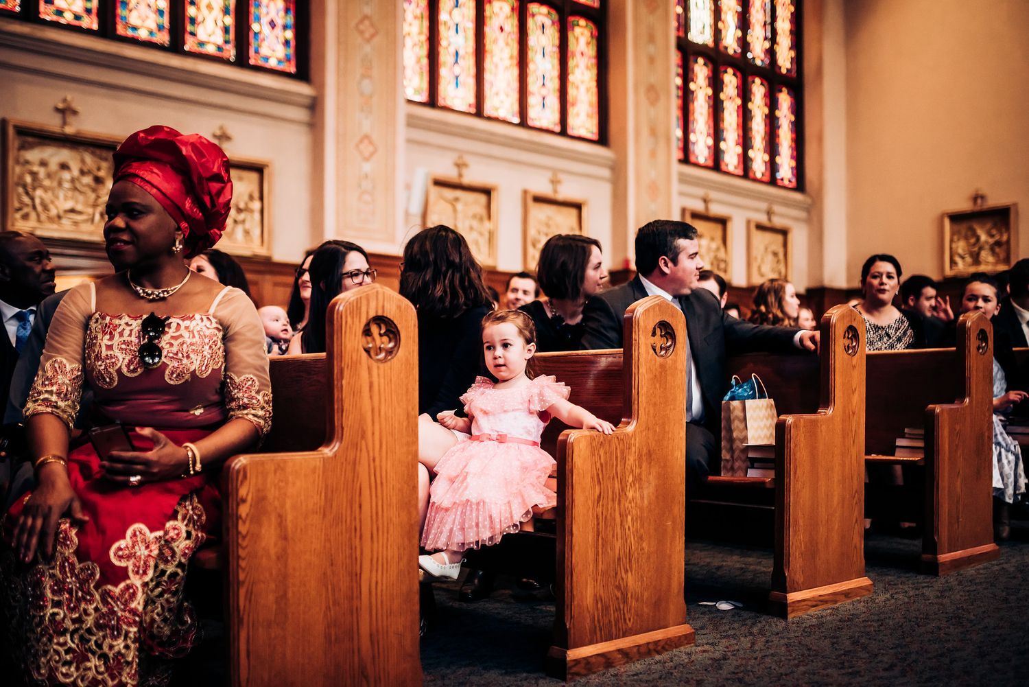 Wedding guests sit in wooden church pews with stained glass windows visible in the background.