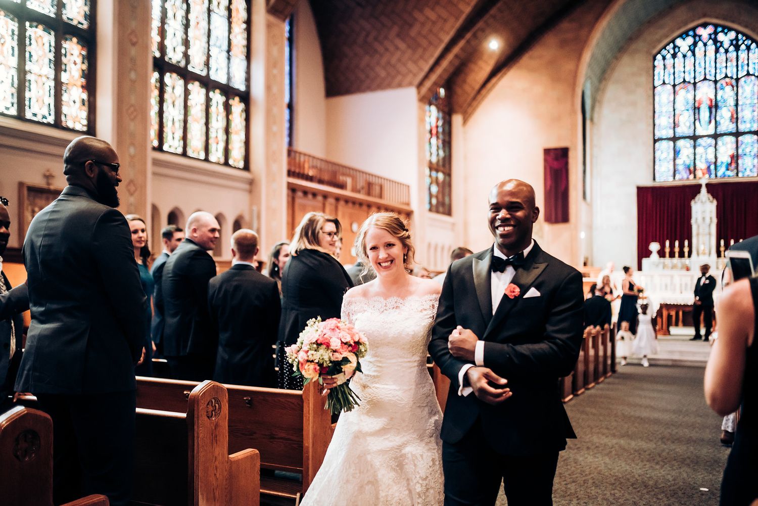 Joyful wedding recessional down church aisle with stained glass windows in background.