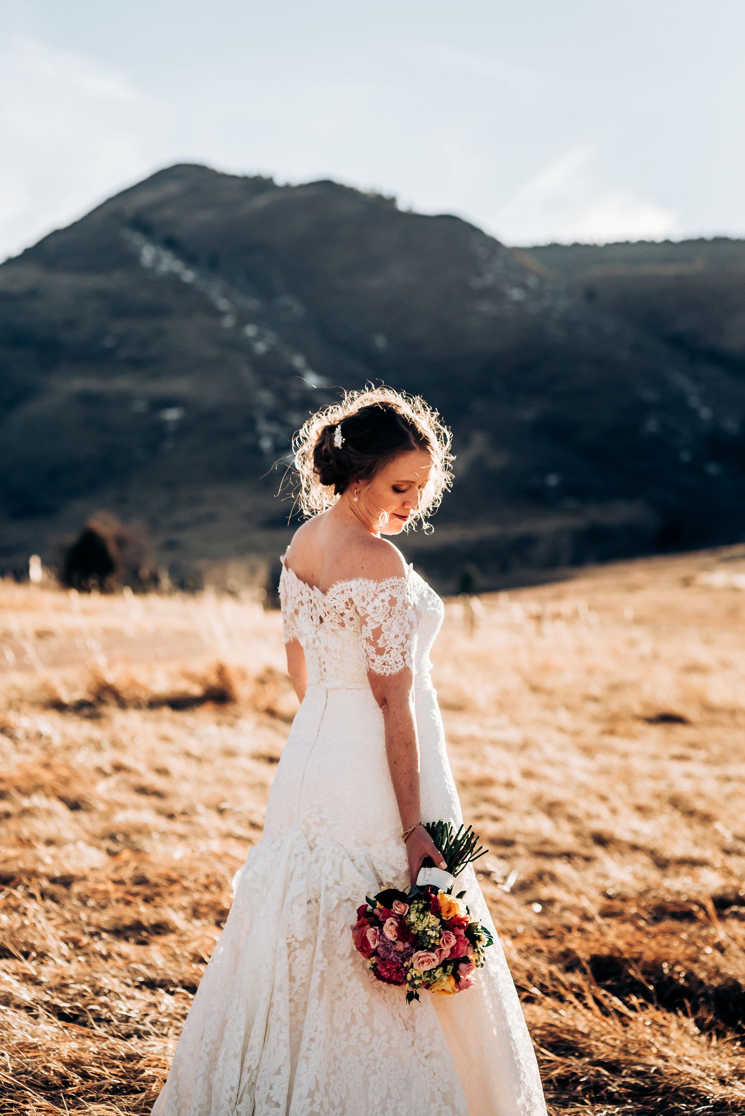 Elegant wedding dress with lace details and floral bouquet photographed against mountain backdrop.