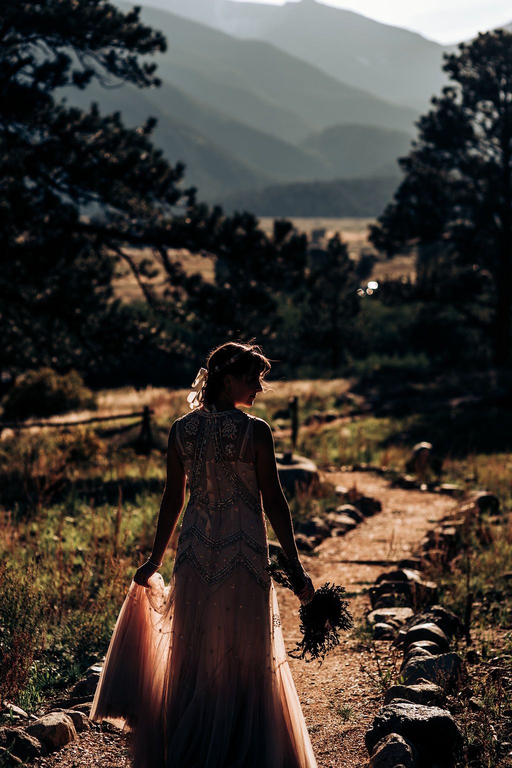 Sunset silhouette of dress flowing on mountain path with layered peaks behind.