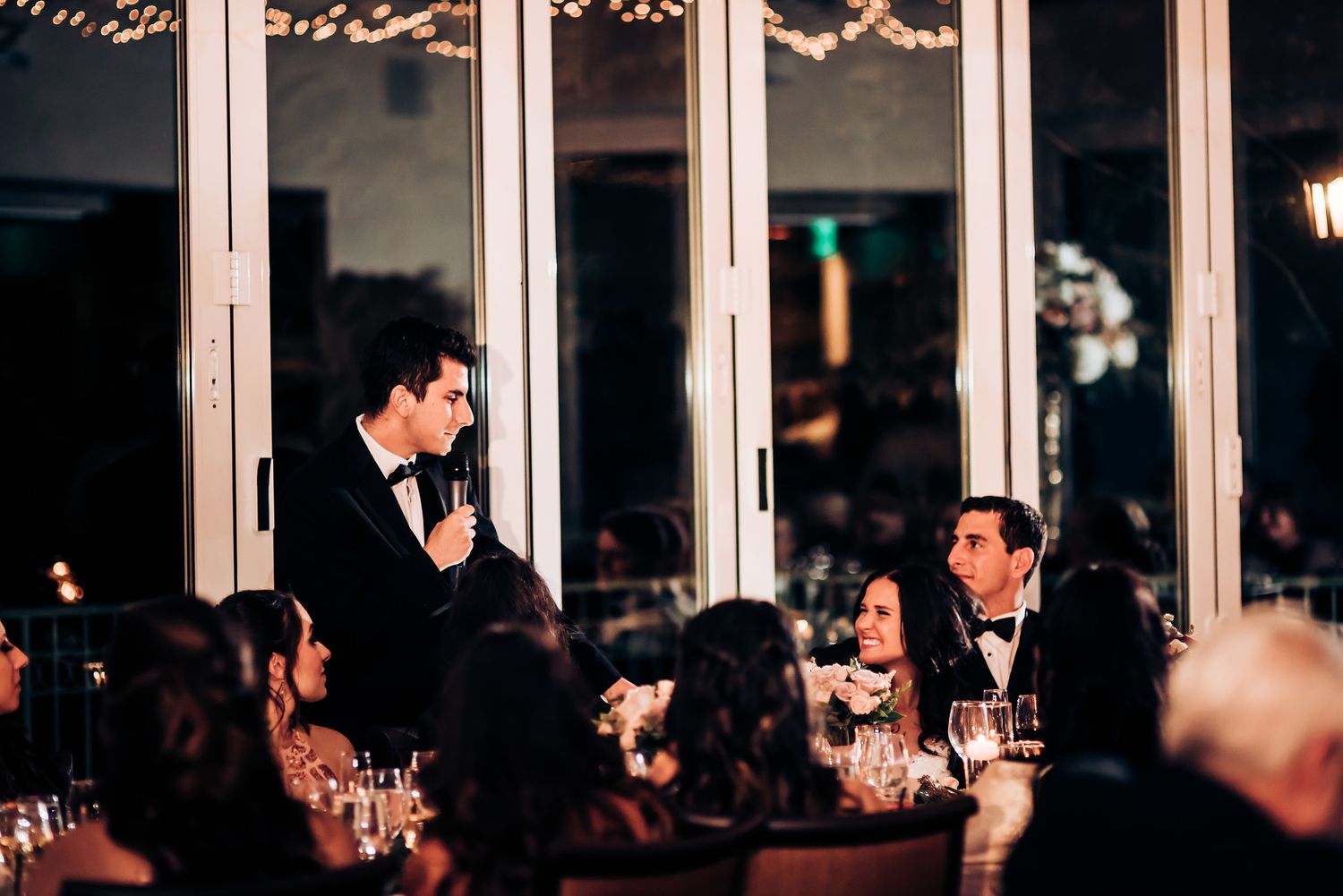 Wedding guests celebrate during a toast at an elegant evening reception with floor-to-ceiling windows.