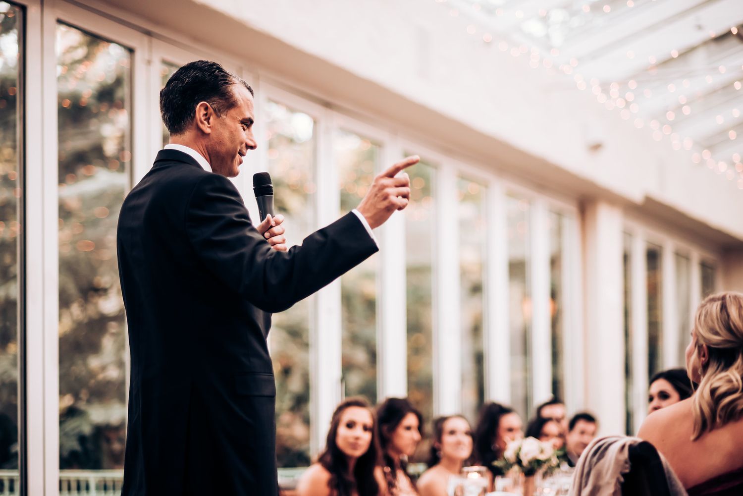 Speaker delivers an animated speech during an elegant wedding reception in a bright venue with glass windows.