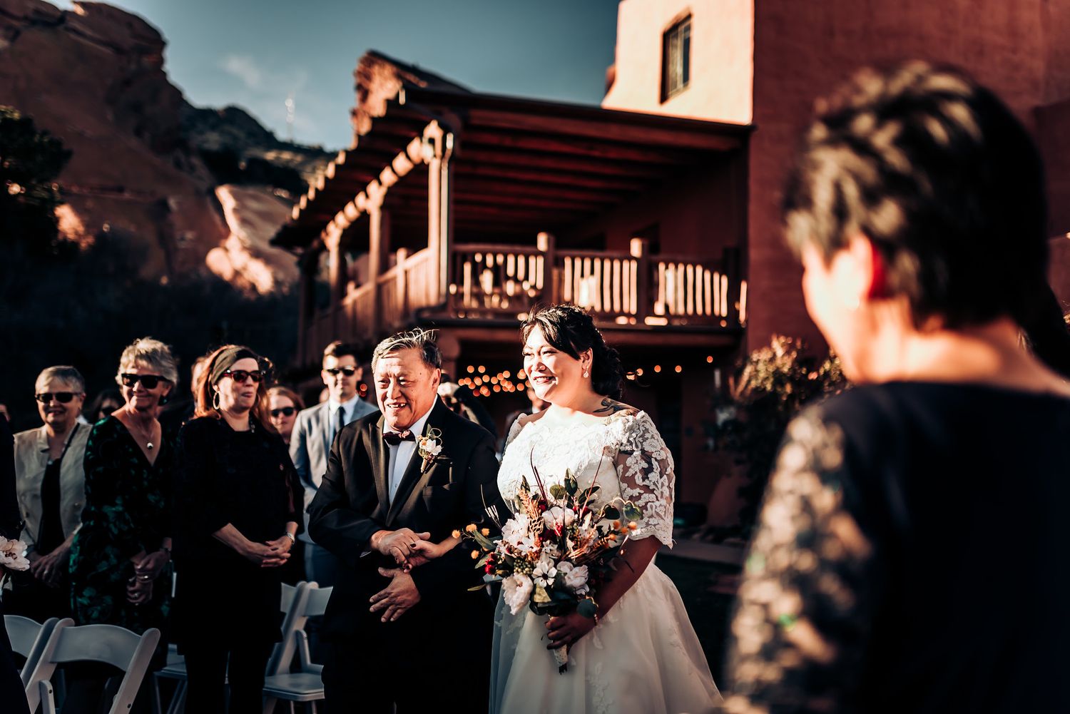 A wedding ceremony takes place at a rustic venue with red rock formations in the background during sunset.