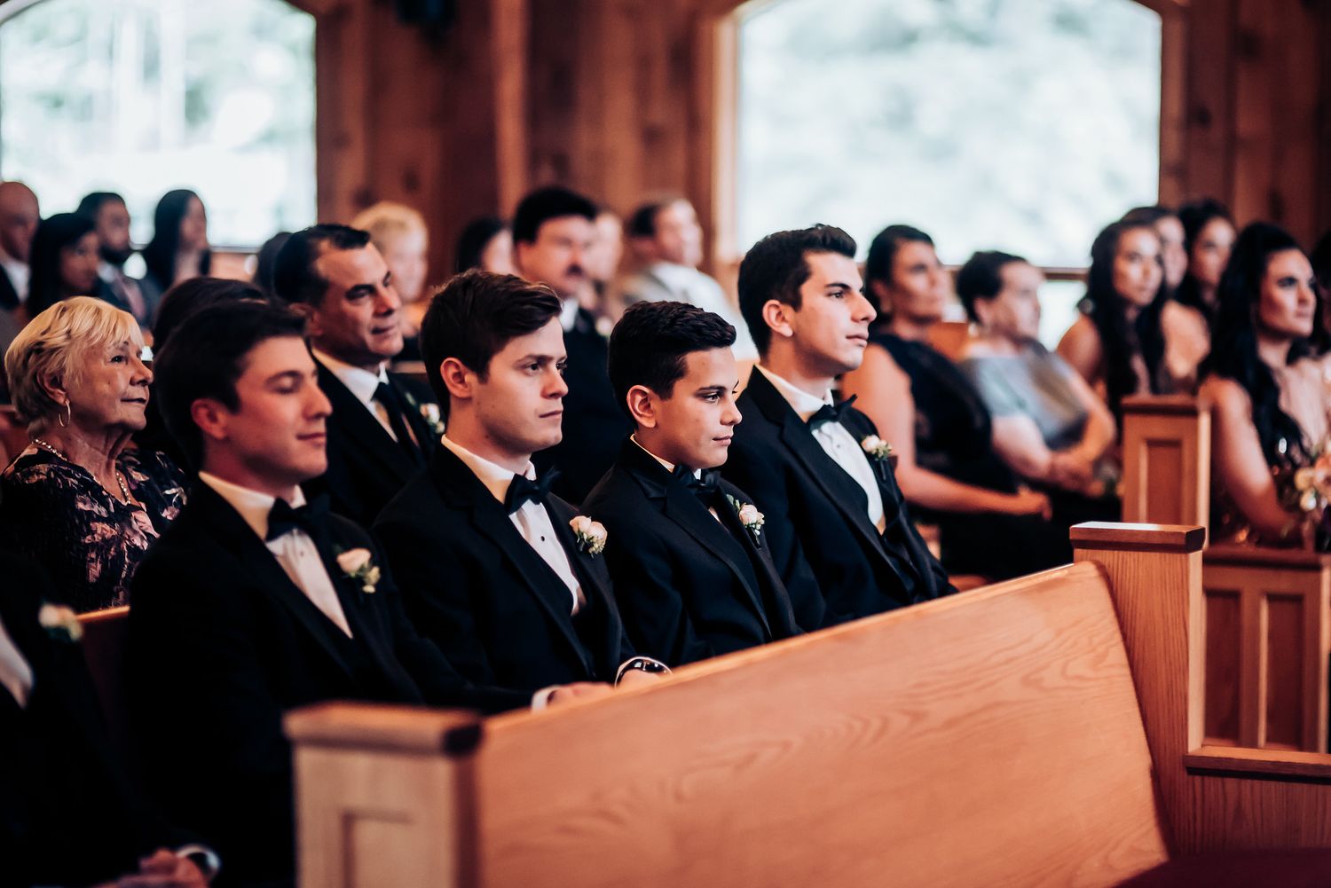 Wedding guests in formal attire seated in wooden church pews.