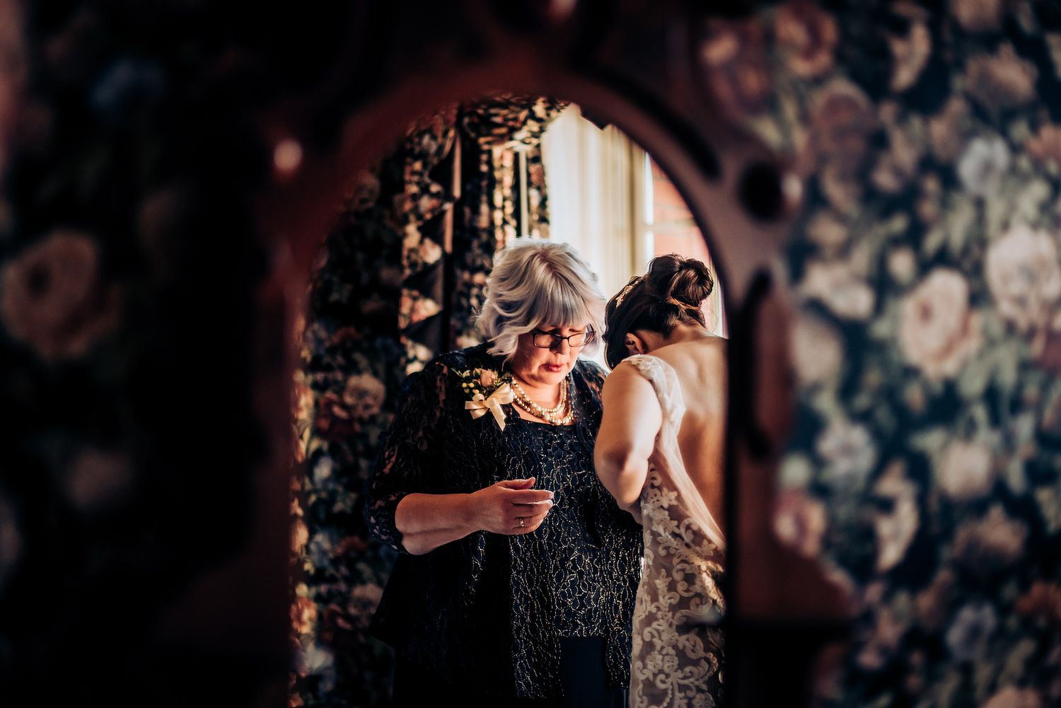 Wedding preparation scene captured through an ornate mirror with floral wallpaper backdrop.