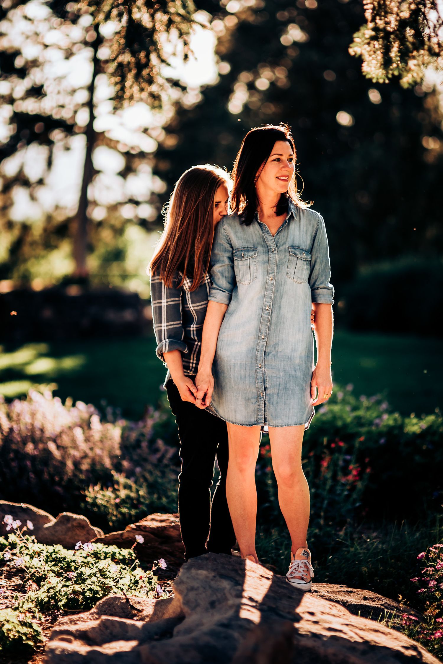Two people stand together on rocky ground with sunlit trees in the background.