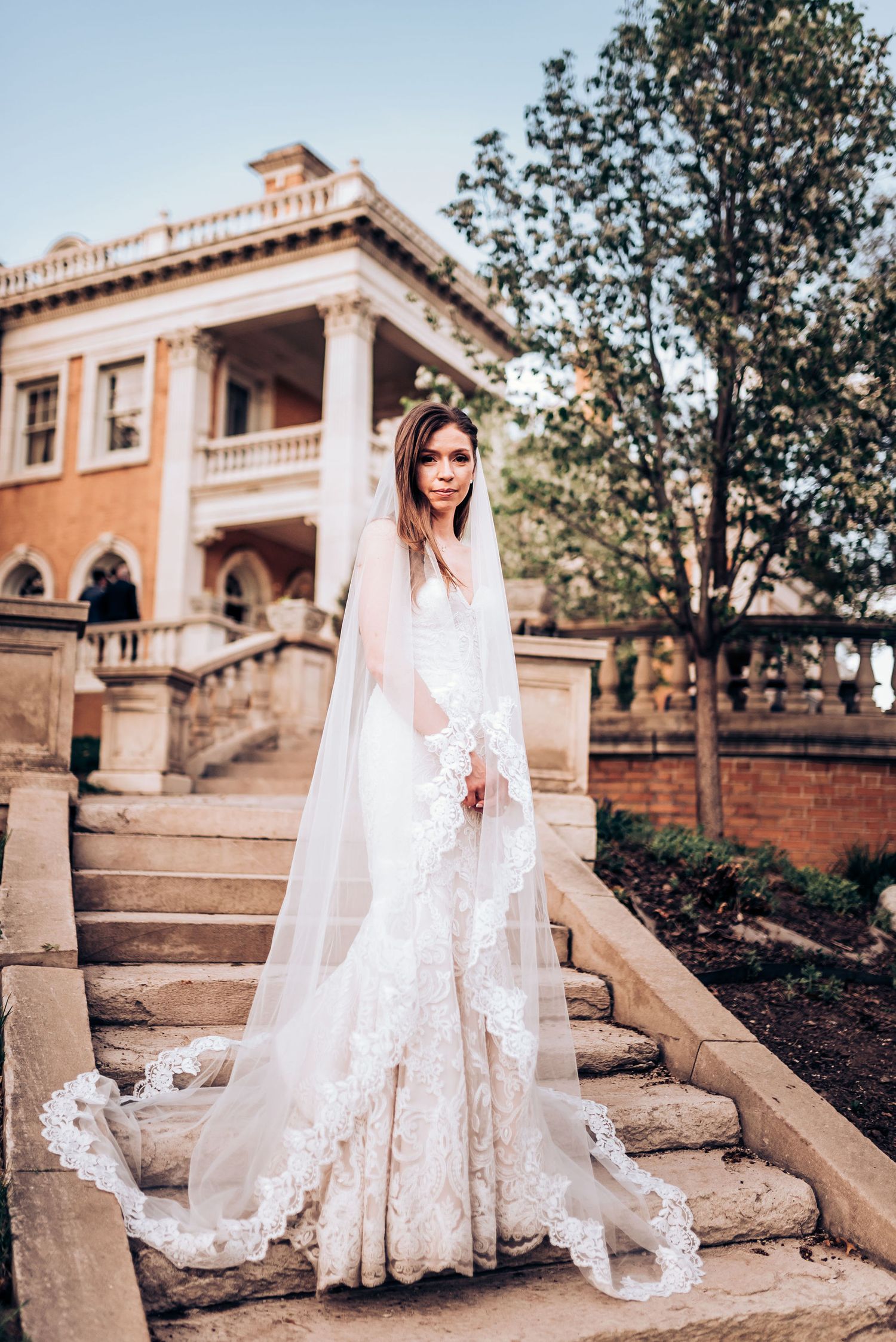 Lace wedding gown trails elegantly on historic stone steps of Mediterranean villa.