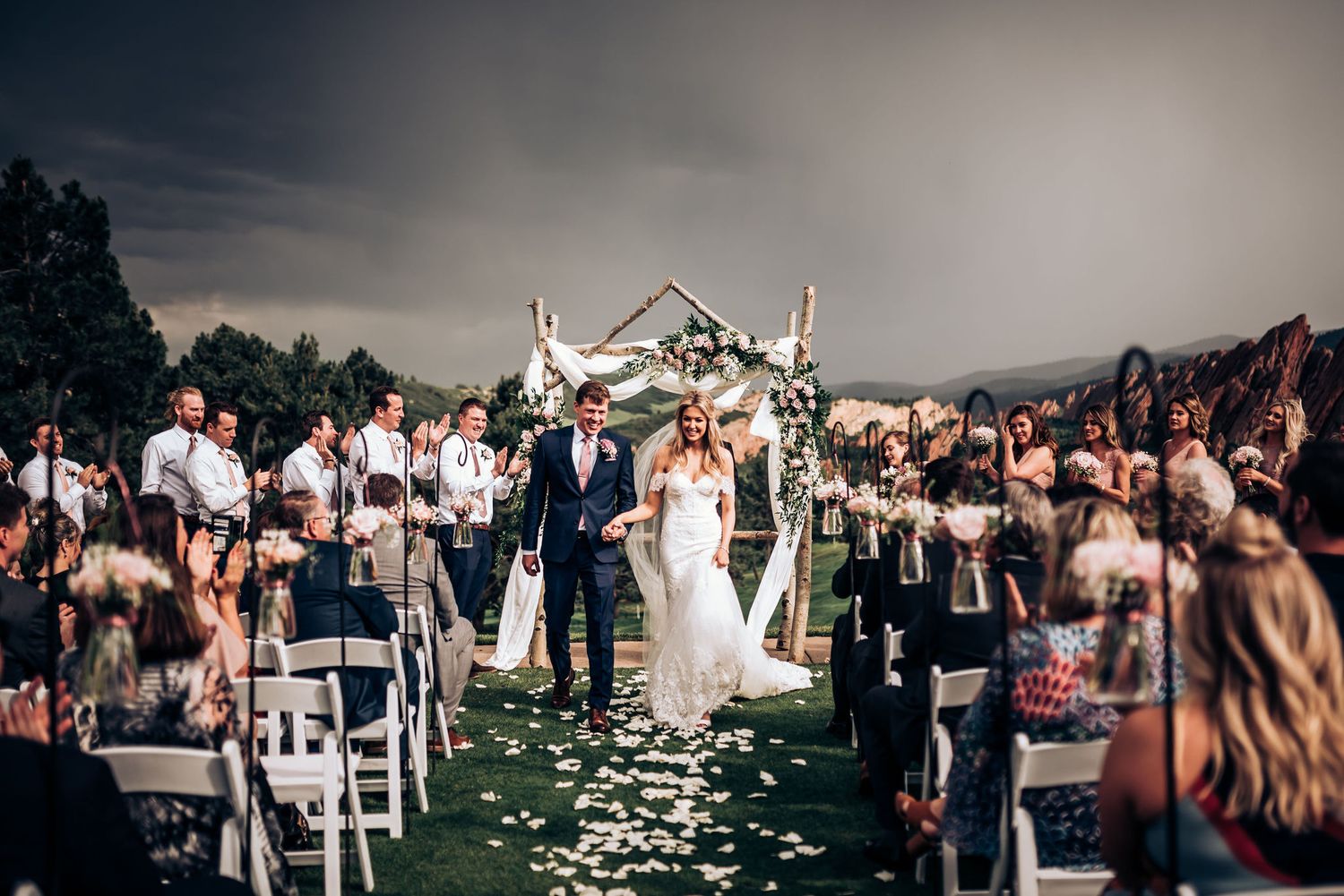 Elegant outdoor wedding ceremony with white floral arch and rose petals scattered down the aisle.