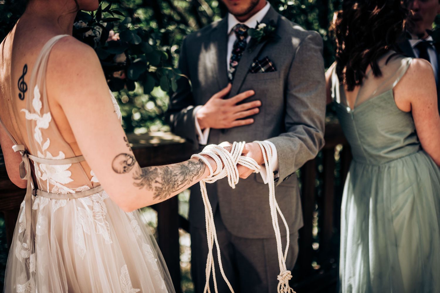 Close-up of wedding ceremony details showing lace dress and grey suit in dappled forest light.