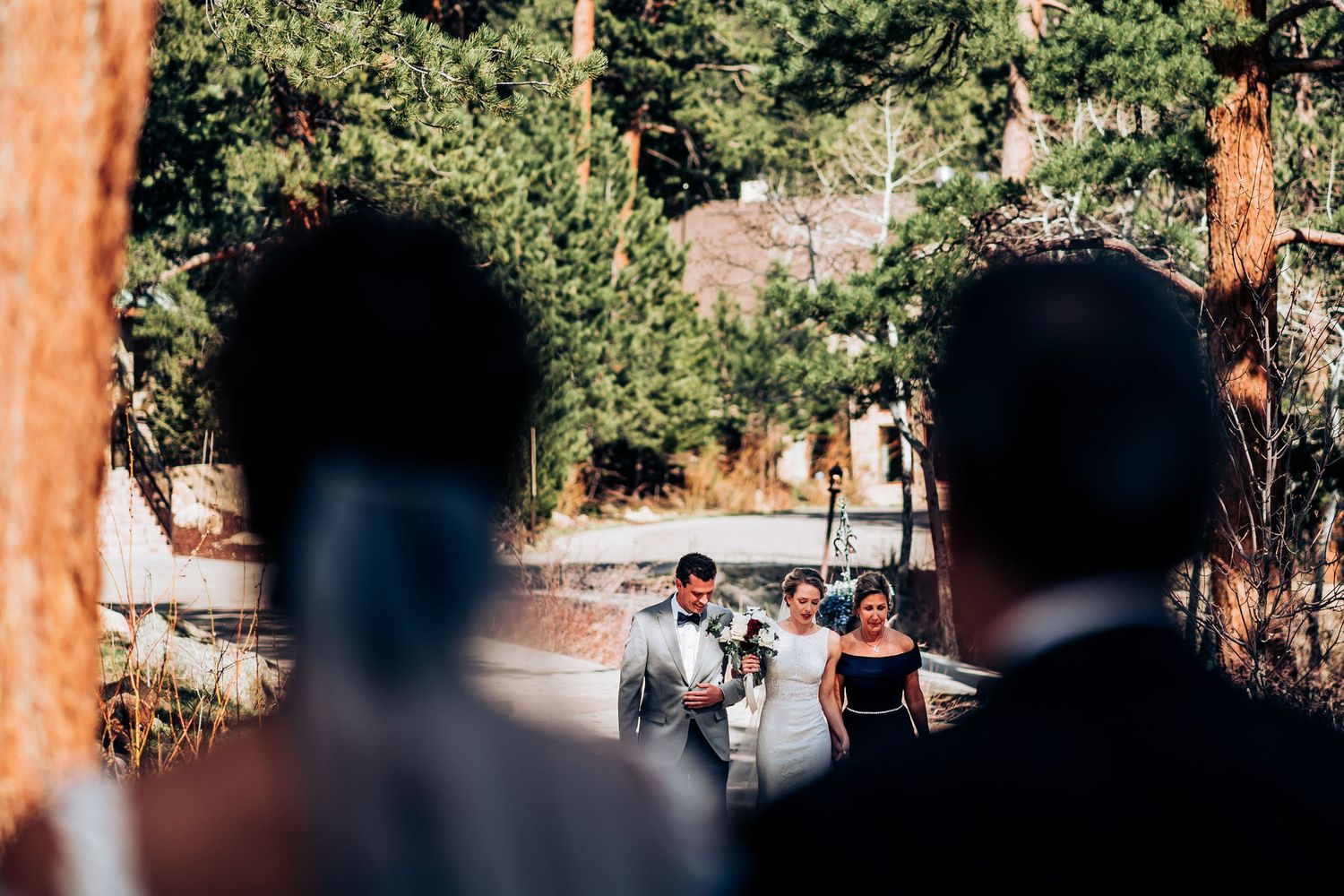 Outdoor wedding ceremony framed through silhouettes of guests in a wooded mountain setting.
