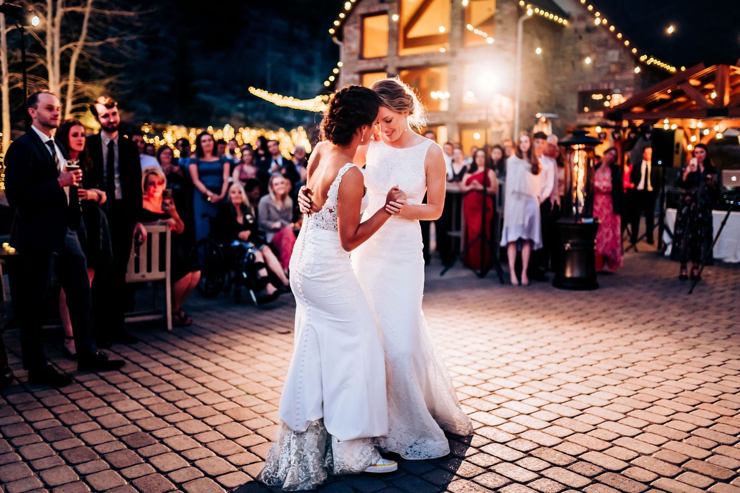 Wedding couple dancing on a brick patio with rustic lodge and string lights in background.