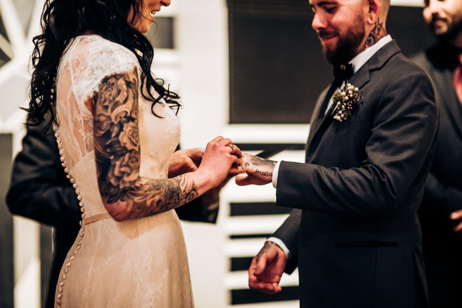 Close-up of ring exchange during wedding ceremony showing tattooed arm and formal attire.