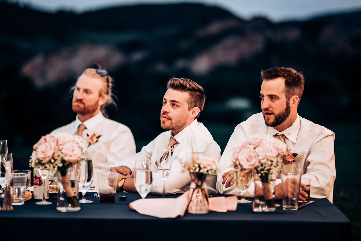 Groomsmen in light colored suits seated at reception table listening to toasts.