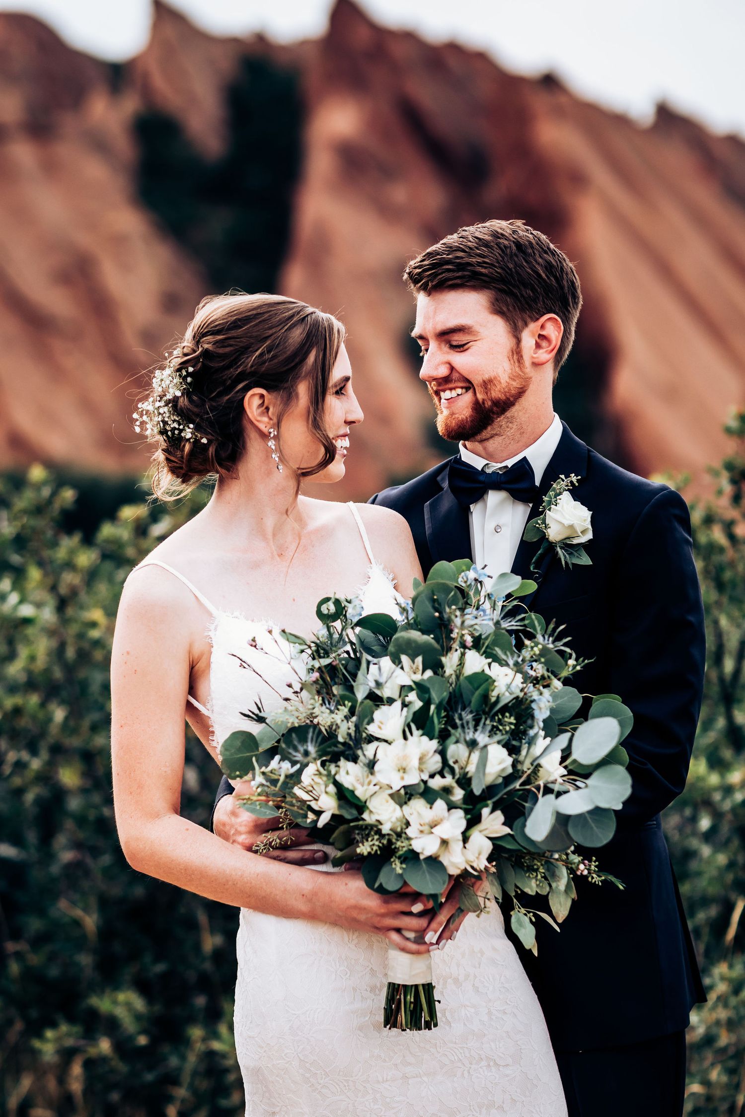 A couple shares a romantic moment while holding a white and green bouquet against red rock formations.