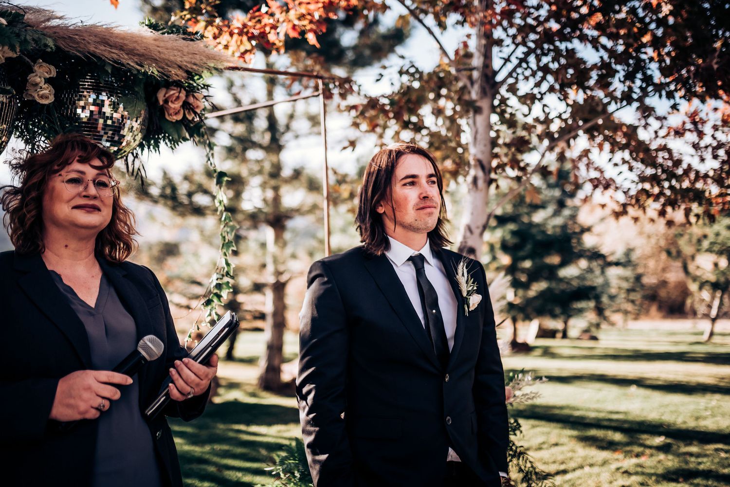 Wedding ceremony participants standing under a floral arch in an autumn outdoor setting.