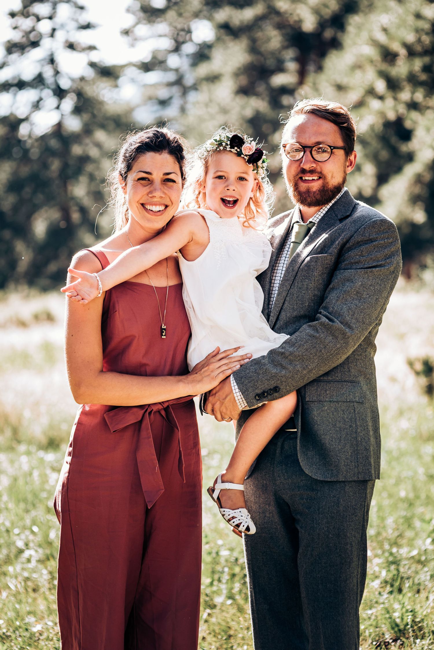 Happy family poses together outdoors in a sunny mountain meadow with pine trees.