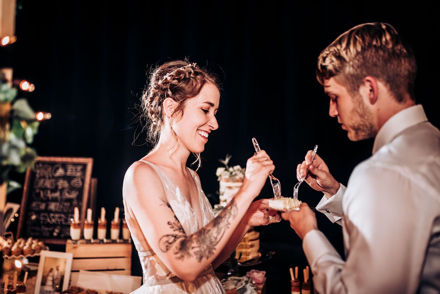 Newlyweds share a playful moment while feeding each other cake at their wedding reception under ambient lighting.