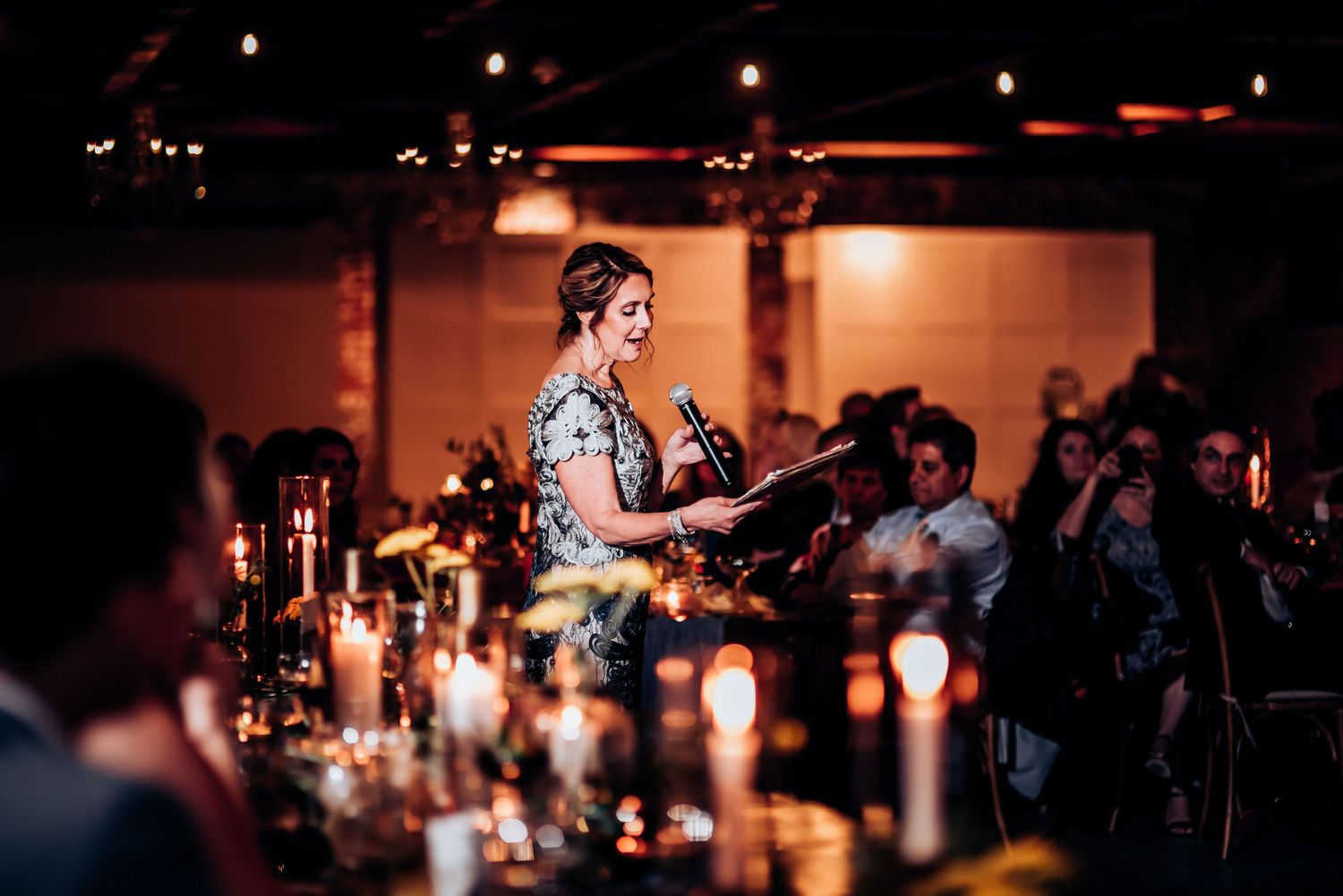 Speaker at evening reception addresses guests among candlelit tables.
