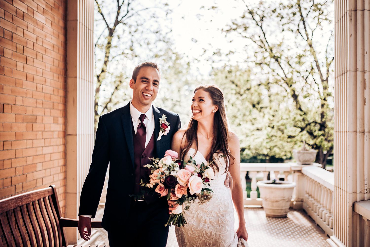 Couple sharing joyful moment on brick columned porch with pink floral bouquet.
