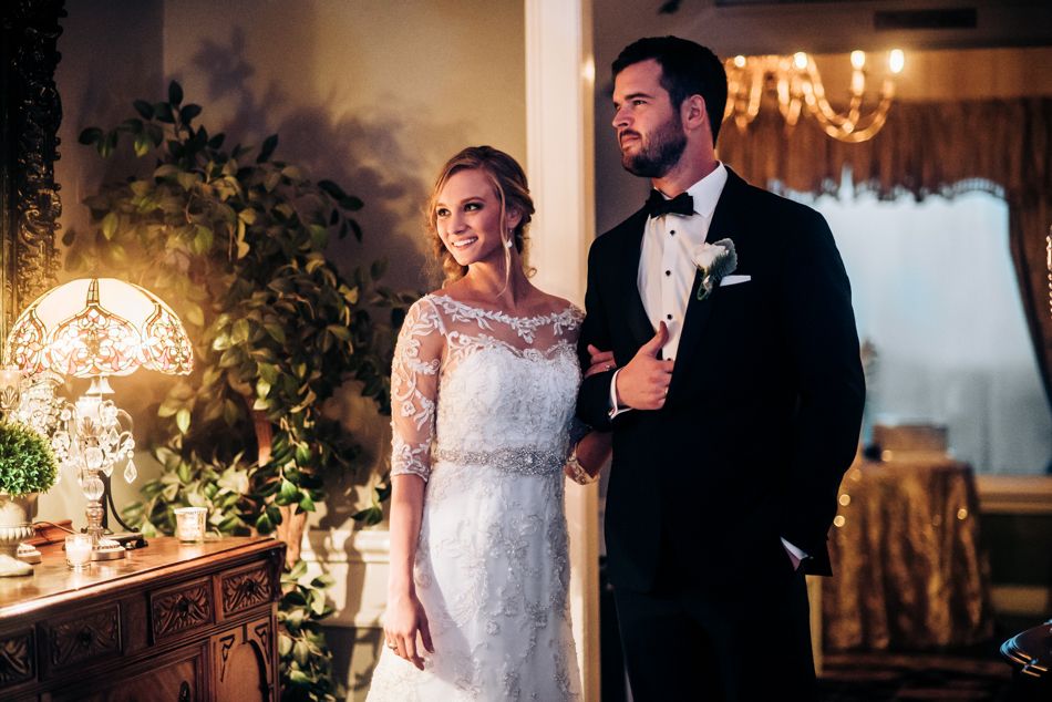 Elegant wedding couple poses beside ornate table with glowing lamp in romantic evening setting.